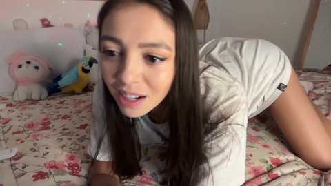 Video of a young woman with long black hair, light skin, and slender physique, wearing a white t-shirt, lying on a floral-patterned bed with stuffed animals.