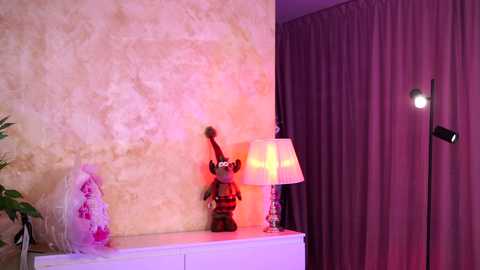 A video of a cozy, warmly-lit room corner featuring a red reindeer figurine, a pink Christmas ornament, and a white lamp on a white dresser against a textured, light beige wall.
