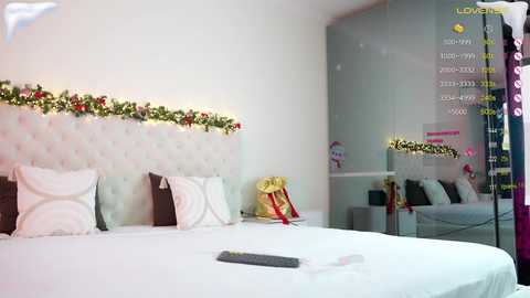 Video of a modern, minimalist bedroom with a white tufted headboard, white bedding, and a festive garland. A mirror reflects the room's decor, including a digital clock and a calendar.