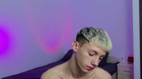 Video of a shirtless, young, white male with short, platinum blonde hair, leaning forward, eyes closed, in a dimly lit bedroom with purple lighting.