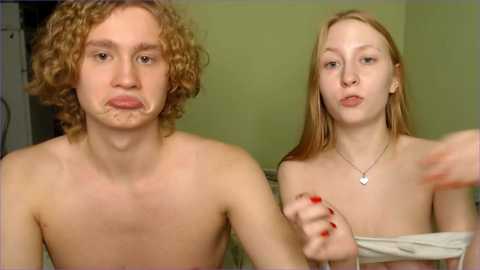 Video of a young, shirtless, curly-haired man with facial makeup, and a topless woman with long, straight hair, both with fair skin, in a dimly lit room.