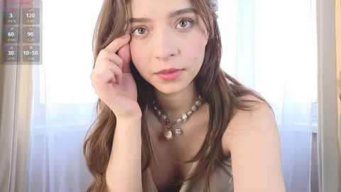 Video of a young woman with long, wavy brown hair, light skin, and green eyes, wearing a beige dress and a beaded necklace, leaning on a window sill with sheer curtains in the background.