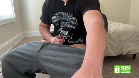 Video of a man with a medium build, light skin, wearing a black \"Gym\" T-shirt and gray sweatpants, sitting on a beige carpeted floor in a simple, dimly lit room.