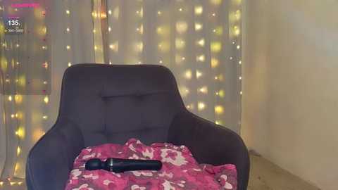 A video of a dark gray armchair with a pink blanket featuring white hearts. A black vibrator lies on the blanket. The background is adorned with golden fairy lights.