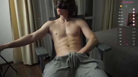 Video of a shirtless, muscular, light-skinned man with short brown hair, wearing a black blindfold, sitting in a chair with grey sweatpants. Background includes a grey couch, wooden floor, and dim lighting.