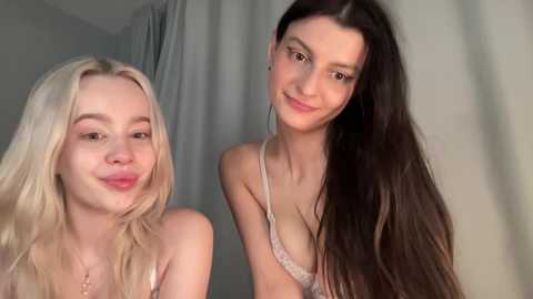 Video of two young women, one with blonde hair and fair skin, the other with long dark hair and olive skin, smiling in lingerie, indoors with neutral background.