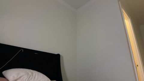 Video of a minimalist bedroom corner with white walls, a black TV on a dark wooden headboard, a white pillow, and a partly open beige door.