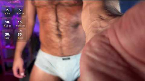Video of a muscular man with a hairy chest wearing white briefs, standing next to another man. The background is blurred, with a digital overlay showing fitness stats.