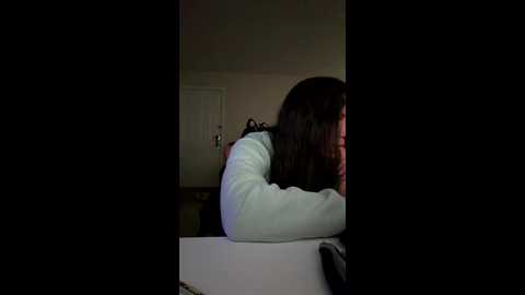 Video of a woman with long, dark hair, wearing a white sweater, leaning over a table, head down, in a dimly lit room with a closed door.