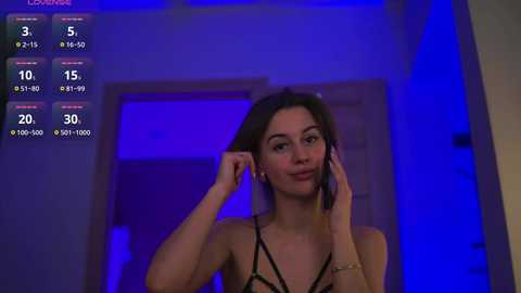 Video of a young woman with long brown hair, wearing black lingerie, adjusting her headphones while standing in a dimly lit room with blue lighting.