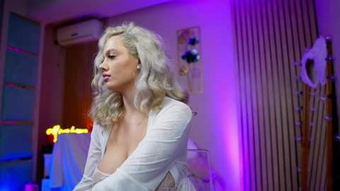Video of a blonde woman in a white, sheer, lace-trimmed robe, standing in a brightly lit, modern room with purple and blue lighting, decorative items, and a door in the background.