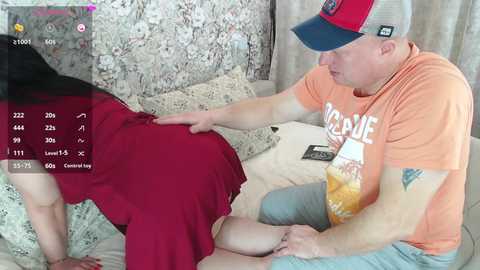 Video of a middle-aged, muscular, fair-skinned man with a tattoo, wearing an orange shirt and baseball cap, touching the back of a woman with long black hair, wearing a red dress, on a bed.