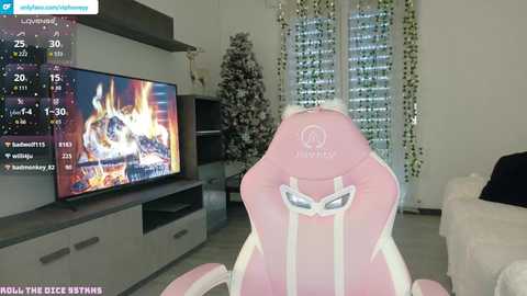 Video of a gamer seated in a pink Razer gaming chair, facing a large flat-screen TV displaying a fire in a snowy forest scene. Background features a white Christmas tree with lights and a modern living room.