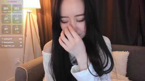 Video of a young Asian woman with long black hair, wearing a white off-shoulder sweater, sitting on a beige couch, covering her mouth with her hand, in a warmly lit room with a lit lamp in the background.