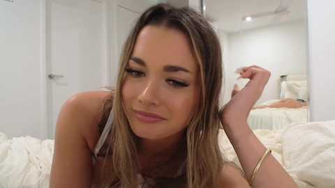 Video of a light-skinned woman with long, wavy brown hair, wearing a sleeveless top, lying on a bed with white sheets, in a modern, minimalist bedroom.