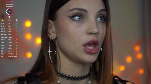 Video of a young woman with fair skin, long brown hair, blue eyes, and a septum piercing. She wears a black choker and large silver earrings. Background has blurred orange lights and a digital calendar.