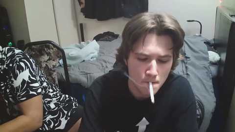 Video of a young man with long brown hair, wearing a black T-shirt, lying on a bed with a white arrow taped to his mouth, surrounded by messy bedding and a patterned chair in a dimly lit room.