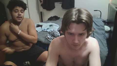 Video of a shirtless, muscular man with curly hair and a thin man with long hair, both in black shorts, in a messy bedroom with clothes scattered around.
