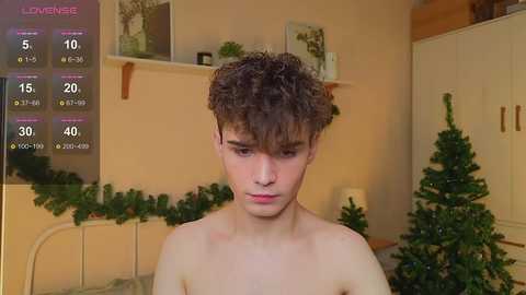 Video of a young, shirtless boy with curly brown hair, fair skin, and a neutral expression, standing in a cozy room with Christmas decorations, a tree, and a bed.