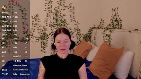 Video of a woman with light skin, dark hair, and a black outfit, seated on a blue couch with orange pillows, surrounded by green ivy vines. A digital screen displays various text and numbers, indicating a live stream or social media broadcast.