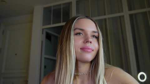 Video of a young woman with long, straight blonde hair, fair skin, and pink lips, wearing a beige top. Background shows a partially open white-framed door and light-colored walls.