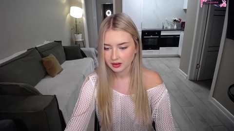 A video of a blonde Caucasian woman with fair skin and a slender physique, wearing a white off-shoulder knitted sweater, sitting in a modern living room with gray sofas, a white kitchen, and a lamp.