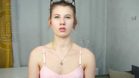 Video of a fair-skinned, slim young woman with blonde hair, wearing a light pink bra, a gold necklace, and sunglasses perched on her head, standing in a modern, minimalist room with grey curtains and a yellow patterned wall.