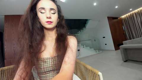 Video of a young woman with long, wavy brown hair, wearing a plaid top, sitting on a chair in a modern, minimalist living room with light-colored walls and a staircase.