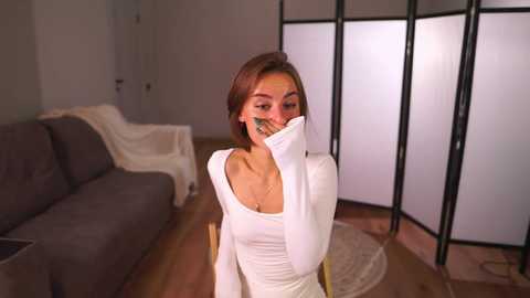 Video of a young woman with short brown hair, wearing a white long-sleeve top, covering her mouth with a hand, sitting on a wooden floor in a modern living room with a gray couch and black folding screen.