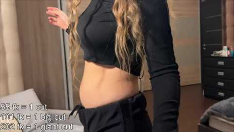 Video of a woman with long, wavy blonde hair in a black crop top, midsection visible, standing in a modern bedroom with beige curtains and dark furniture.