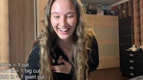 Video of a smiling young woman with long, wavy, blonde hair, wearing a black top, standing in a cluttered room with wooden furniture and cabinets.