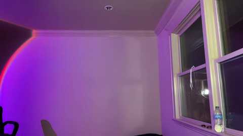 Video of a dimly lit, minimalist room with a large window showing a cityscape, a small chair, and a water bottle on a table, illuminated by soft purple and red lighting.
