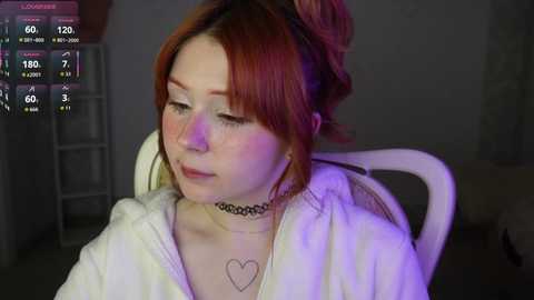 Video of a fair-skinned woman with red hair, wearing a choker necklace and a white robe, seated indoors with purple lighting, displaying VR headset.