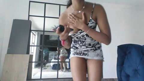 Video of a young woman with medium skin tone, wearing a graphic black-and-white tank top, standing in front of a large mirror in a modern, minimalist room.