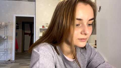 Media: Video of a young Caucasian woman with light brown hair, wearing a gray shirt, sitting in a home with beige walls, a white door, and a wooden floor.