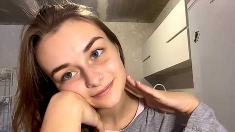 Video of a young woman with fair skin, brown hair, and blue eyes, resting her chin on her hand, wearing a gray shirt in a modern, minimalist kitchen with white cabinets.