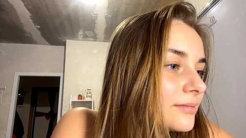 Video of a young woman with light skin and long, straight, blonde hair, looking contemplative. She has a slender build and is indoors, possibly in a bathroom or bedroom, with a dimly lit ceiling.