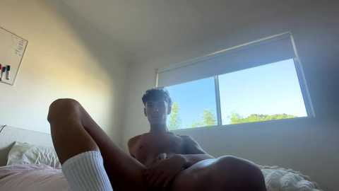 A video of a nude young man with short hair, lying on a bed with white sheets, wearing only white socks, looking out a window at a bright blue sky.
