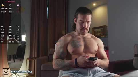Video of a shirtless, muscular man with long hair, tattoos, and a black bracelet, focused on his smartphone in a modern living room.