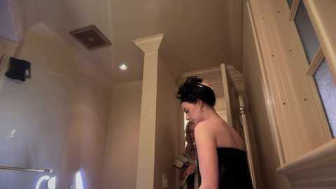 Video of a woman with dark hair styled in an updo, wearing a strapless black dress, standing in a beige-tiled bathroom with white trim and a shower curtain.