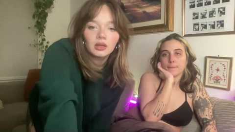 Video of two young women, one with straight brown hair, wearing a green sweater, and the other with wavy blonde hair, black bra, leaning on a bed. Background includes a framed picture and plants.