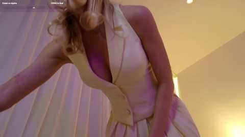 Media: A video of a blonde woman in a low-cut white dress, bending forward, with a blurred background of white curtains and a warm yellow light source.