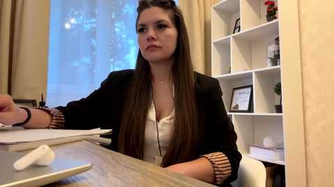 Video of a serious-looking woman with long brown hair in a black blazer and white blouse, sitting at a wooden desk in a modern office.