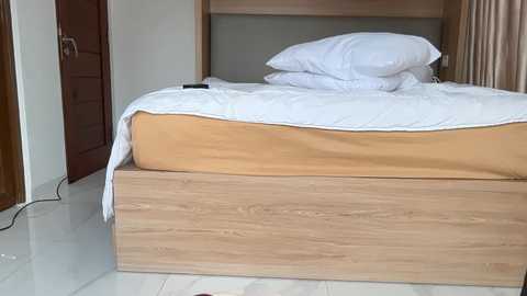 Video of a neatly made bed with white pillows, brown sheets, and a wooden headboard, set against a beige wall and light wooden floor. A black smartphone lies on the bed.