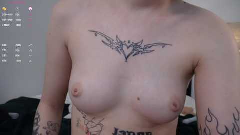 Video of a topless light-skinned person with small breasts, displaying a heart-shaped tattoo on their chest, and various other tattoos on their arms. Background shows a blurred room with a bed and light gray walls.
