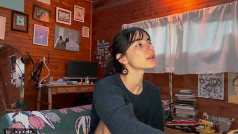 Video of a young woman with dark hair, wearing a black sweater, sitting in a cozy, cluttered room with wooden walls, framed art, and a desk with a computer.