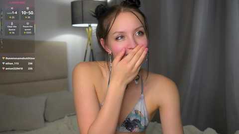Video of a young woman with fair skin, brown hair in buns, wearing a floral halter top, covering her mouth, in a dimly-lit bedroom.
