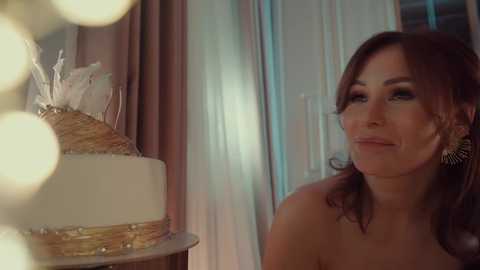 Video of a smiling, middle-aged woman with wavy brown hair, wearing gold earrings, standing next to a two-tiered wedding cake with gold accents and white icing. Soft, warm lighting and bokeh effect.