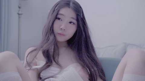 Video of an East Asian woman with long, wavy brown hair, wearing a sheer white lingerie top, sitting on a bed with light blue sheets, in a softly lit room.