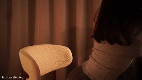 A video of a woman in a cream sweater, seated on a beige, ribbed-textured chair with a curved back, in a dimly lit room with beige curtains.
